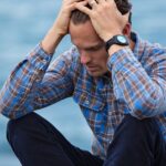 A man in a plaid shirt sits by the water looking distressed, symbolizing stress.